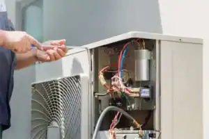 %Focuskw% | John Nugent &Amp; Sons A person uses a screwdriver to repair or maintain an outdoor air conditioning unit with exposed wires and components.