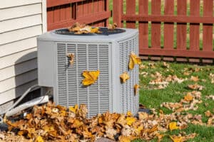 An outdoor air conditioning unit is surrounded by and covered with fallen autumn leaves next to a house and a wooden fence.