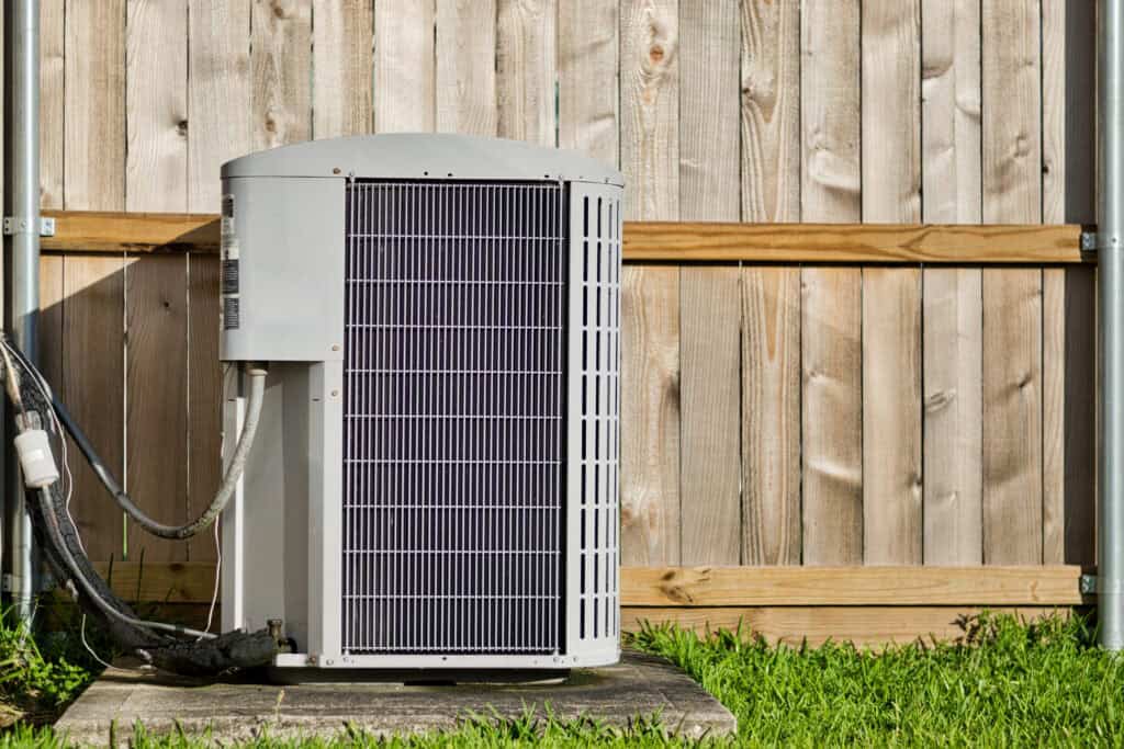 Outdoor air conditioning unit installed on a concrete pad next to a wooden fence, with green grass in the foreground.