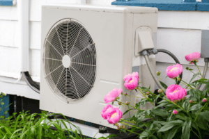 %Focuskw% | John Nugent &Amp; Sons An outdoor air conditioning unit is mounted on the side of a house with blooming pink flowers nearby.