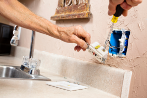 Person installing or repairing an electrical outlet above a kitchen counter, using a screwdriver, with the outlet cover and screws placed nearby.
