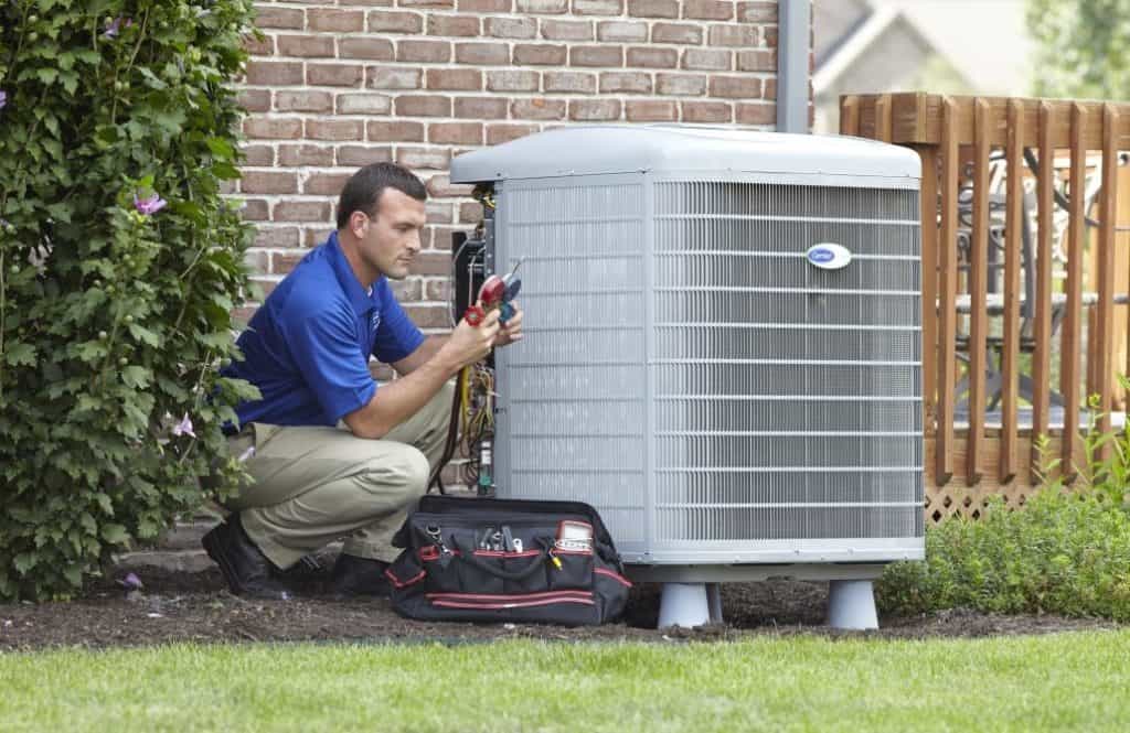 A technician in a blue shirt kneels beside an outdoor air conditioning unit, using tools, with a tool bag on the ground nearby.