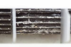Close-up of dirty, dust-covered air conditioner or heater vent behind plastic grilles, showing heavy buildup of debris and grime on metal fins.