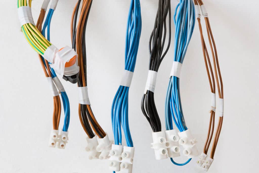 Several bundles of electrical wires, color-coded brown, blue, black, and green-yellow, are grouped and secured with zip ties, ending in white connector blocks against a white wall.