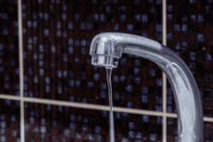 A close-up of a chrome faucet with a thin stream of water flowing, set against a dark tiled background with light grout lines.