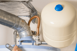 A person’s hand uses a wrench to adjust a fitting on a water heater, which is connected to a metal vent pipe and a beige expansion tank.