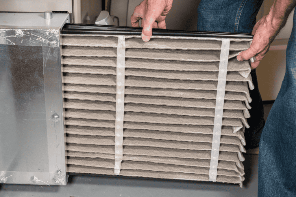 Person sliding a dirty, dust-covered HVAC air filter out of a furnace slot.