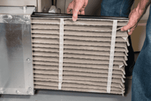 Person sliding a dirty, dust-covered HVAC air filter out of a furnace slot.